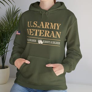 Op de afbeelding: Een groen hoodie met de tekst "U.S. ARMY VETERAN" in gouden letters. De tekst "Once a soldier, always a soldier" staat onder de hoofdtekst. Een kleine Amerikaanse vlag bevindt zich op de linkermouw.