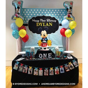 K&ouml;nnte beinhalten: Dekoration f&uuml;r einen ersten Geburtstag im Mickey Mouse-Stil. Enth&auml;lt einen Kuchen, Luftballons und ein Banner mit Fotos. Der Hintergrund zeigt die Aufschrift "Happy First Birthday Dylan."