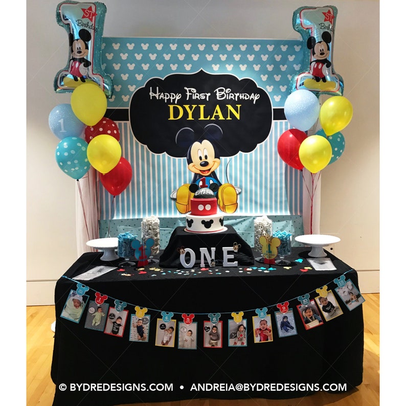 K&ouml;nnte beinhalten: Dekoration f&uuml;r einen ersten Geburtstag im Mickey Mouse-Stil. Enth&auml;lt einen Kuchen, Luftballons und ein Banner mit Fotos. Der Hintergrund zeigt die Aufschrift "Happy First Birthday Dylan."