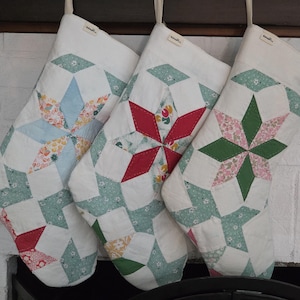 May include: Three quilted Christmas stockings featuring a star pattern made from various floral and solid fabric patches in shades of light blue, red, green, and white. Each stocking has a plain white cuff with a small rectangular brand label.