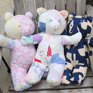 May include: Three handmade patchwork teddy bears in pastel colors sit on a wooden chair. The bears have different patterns, including floral, polka dots, and geometric shapes. Each bear has a unique color scheme, with pink, blue, and white dominating.