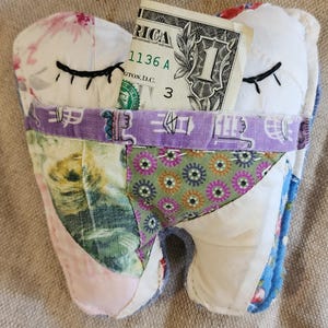 May include: A handmade tooth-shaped fabric pouch with a dollar bill peeking out. The pouch is made of various patterned fabrics, including floral and geometric designs, with embroidered closed eyes. The dollar bill shows the text "1" and "America".