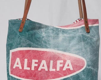 Puede incluir: Una bolsa de lona de color verde azulado y blanco con asas de cuero marrón. La bolsa tiene un óvalo rojo con la palabra "ALFALFA" impresa en blanco. La parte inferior de la bolsa tiene el texto "GRIMM ALFALFA" impreso en negro.