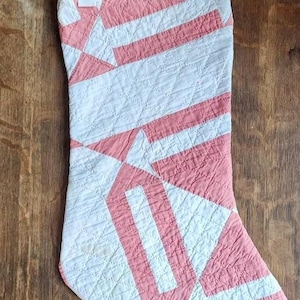 May include: A quilted Christmas stocking with a geometric pattern of white and coral-colored stripes. The stocking has a loop for hanging and is made of fabric. The design is a patchwork style, with the fabric pieces sewn together.