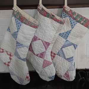 May include: Three quilted Christmas stockings hanging in a row. Each stocking features a patchwork design with white fabric panels interspersed with blue, red, and pink patterned squares. The cuffs are decorated with a red and green floral print.