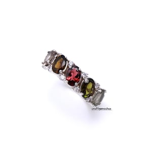 May include: A silver ring featuring a row of oval-cut gemstones in various colors, including brown, red, and green, interspersed with smaller white stones. The ring is set against a white background.