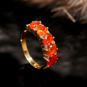 May include: A gold ring with a series of oval-cut, vibrant orange gemstones. Tiny, clear accent stones are set between the larger gems. The ring's band is a polished gold colour, creating an elegant and striking piece of jewellery.