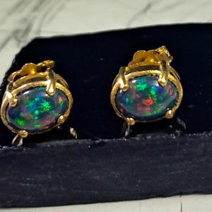 Puede incluir: Un par de pendientes de botón dorados, cada uno con una ópalo ovalado oscuro con destellos verdes, azules y rojos. Los ópalos están engastados en un bisel dorado con detalles decorativos.