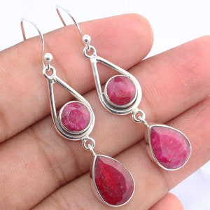 May include: Silver dangle earrings featuring three ruby gemstones. Each earring has a round ruby set within a teardrop-shaped frame, with a pear-shaped ruby dangling below. The rubies are a vibrant red color.