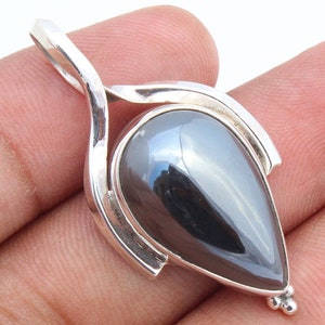 May include: A teardrop-shaped pendant with a polished, dark gray stone set in a silver metal frame. The pendant has a loop at the top for a chain and a small cluster of three beads at the bottom.