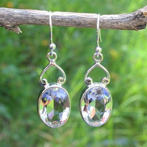 Puede incluir: Un par de pendientes colgantes plateados con piedras preciosas ovaladas, facetadas y transparentes. Los pendientes cuelgan de un diseño curvo y un gancho simple. El fondo es verde borroso.