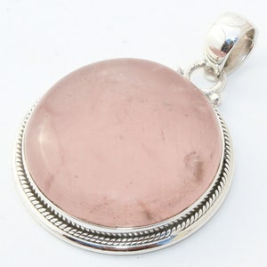 May include: A round pendant featuring a large, polished rose quartz stone. The stone has a soft pink hue and is set in a silver-colored metal frame with a decorative rope-like border. The pendant includes a silver bail for a chain.