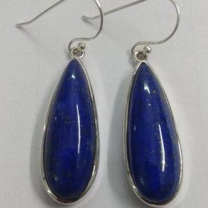 May include: A pair of teardrop-shaped earrings featuring deep blue lapis lazuli stones set in silver-colored metal. The earrings have a polished finish and a simple, elegant design with a hook closure. The stones have a speckled appearance.