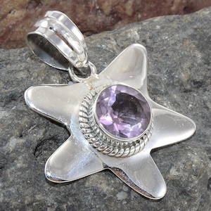 May include: A silver-toned pendant in the shape of a star, featuring a round, light purple gemstone at its center. The gemstone is encircled by a decorative, twisted metal border. The pendant has a loop for a chain.