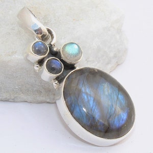 May include: A silver pendant featuring an oval labradorite gemstone with blue and gray hues. The pendant also includes three smaller, round gemstones in shades of blue and green. The jewelry is set against a white background.