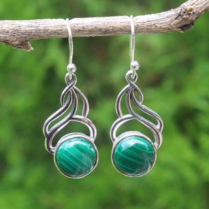 May include: A pair of silver dangle earrings featuring a round, polished green malachite stone. The earrings have a decorative silver swirl design above the stone and a simple hook closure. The background is a soft green.