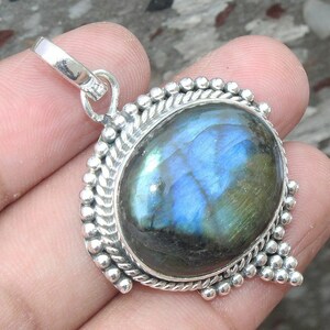 Puede incluir: Colgante ovalado de labradorita con engaste de plata. La piedra preciosa muestra tonos iridiscentes azules y verdes. El colgante tiene un borde decorativo de pequeñas cuentas de plata y un lazo para una cadena.