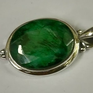 Puede incluir: Colgante ovalado con una piedra preciosa de esmeralda facetada engastada en un bisel de plata. La esmeralda es de color verde intenso. El colgante incluye una fianza de plata para una cadena.