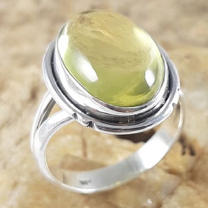 Puede incluir: Un anillo de plata con una gema grande, ovalada y de color amarillo pálido. La gema está engastada en un bisel y el anillo tiene un diseño de caña dividida. El anillo se muestra sobre una superficie marrón claro.