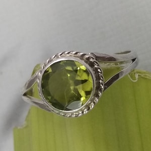 Puede incluir: Un anillo de plata con una piedra preciosa grande, redonda y verde claro. La piedra preciosa está engastada en un bisel con un diseño de cuerda trenzada. La banda del anillo se divide en dos hebras delicadas.