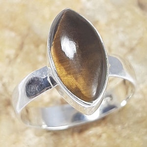 Puede incluir: Un anillo de plata con una piedra ojo de tigre en forma de marquesa. La piedra es de color marrón cálido con vetas doradas, engastada en una banda de plata pulida. El diseño del anillo es sencillo y elegante.