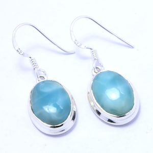 May include: A pair of silver drop earrings featuring oval, light blue gemstones. The gemstones are set in a silver bezel and have a polished, glossy finish. The earrings have a simple, elegant design.