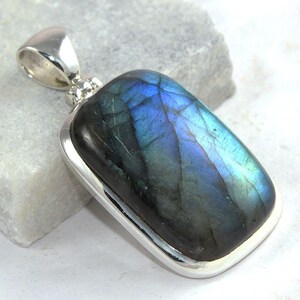 May include: A rectangular labradorite pendant with a silver setting. The stone displays iridescent blue and green hues. The pendant has a silver bail for attaching to a chain. The gemstone is approximately 1.5 inches long.