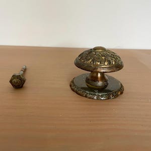 Puede incluir: Antiguo pomo de puerta de latón con un diseño floral ornamentado. El pomo tiene una parte superior en forma de hongo y una base decorativa. También se ve un tornillo separado. El herraje es probablemente para un armario o una puerta.