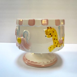 May include: A white ceramic bowl with a pink and white striped rim. The bowl features a carousel scene with a pink horse, a yellow giraffe, and a blue and white striped pole. The bowl has a pink and white base.