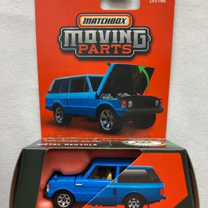 May include: A blue 1975 Range Rover Matchbox toy car with the hood open, displayed on a red and black cardboard backing. The packaging reads "Moving Parts" and "Metal Recyclable".