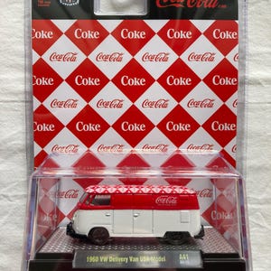 May include: A red and white 1960 Volkswagen Delivery Van die-cast model with Coca-Cola branding. The van is on a black and silver display base with a checkered pattern and the text "1960 VW Delivery Van USA Model" and "A41 24-15".