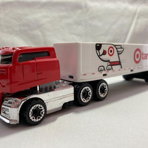 May include: A red and silver toy semi-truck with a white trailer. The trailer features the Target logo and mascot. The truck has black wheels and is set against a white background.