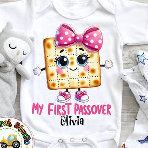 May include: White baby onesie with a cartoon matzah wearing a pink bow and the text "My First Passover Olivia".