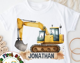 Puede incluir: Camiseta blanca con una ilustración de una excavadora amarilla y el nombre "JONATHAN" impreso en una pancarta marrón. La camiseta tiene un pequeño logotipo con una excavadora de juguete amarilla y el texto "Kids First Studio Things Kids Love".