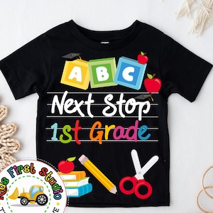 May include: Black t-shirt featuring a colourful ABC block design, a red apple, a pencil, scissors, and the text "Next Stop 1st Grade".