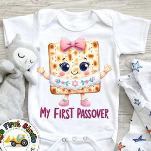 May include: A white baby grow with a cartoon matzah wearing a pink bow and a white sash with a Star of David. The grow has the text "My First Passover" printed on it.