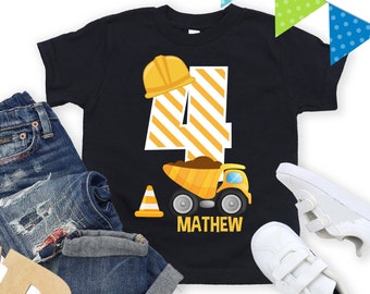 May include: A black t-shirt with a yellow number four and a construction truck graphic. The text "MATHEW" is printed below the truck. The shirt is on a white background with blue jeans, wooden letters, and a gift box.