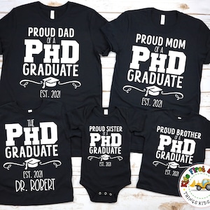 May include: Black t-shirts with white text that reads "Proud Dad of a PhD Graduate Est. 2021", "Proud Mom of a PhD Graduate Est. 2021", "The PhD Graduate Est. 2021 Dr. Robert", "Proud Sister of a PhD Graduate Est. 2021", and "Proud Brother of a PhD Graduate Est. 2021".