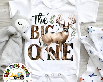 May include: White baby onesie featuring a brown and white deer, green leaves, and the text "The Big One" in a camouflage pattern.