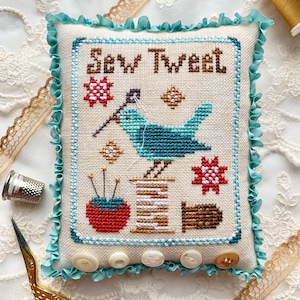 May include: A cross-stitch cushion featuring a blue bird, spools of thread, and a needle and thread. The cushion is edged with a light blue frill and has white buttons around the border. The cushion reads "Sew Tweet".