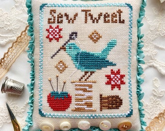 May include: A cross-stitch pillow with a blue bird, spools of thread, and a needle and thread. The pillow is surrounded by a light blue ruffle and has white buttons around the edge. The pillow reads "Sew Tweet".