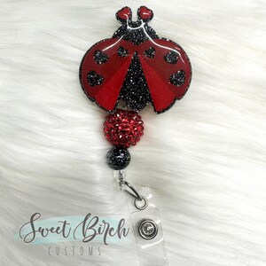 May include: A red and black ladybug charm with glitter and rhinestones, attached to a clear badge holder with a silver clip. The charm is shaped like a ladybug with black spots and a red body. The badge holder has a clear plastic backing and a silver metal clip.