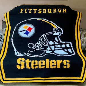 May include: A black and yellow crocheted blanket featuring the Pittsburgh Steelers logo. The logo depicts a football helmet in the team's colours with the words "Pittsburgh Steelers".