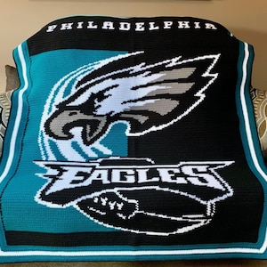 Op de afbeelding: Een gehaakte deken met het logo van de Philadelphia Eagles. De deken is groen, zwart en wit met de naam van het team en een gestileerde adelaar.