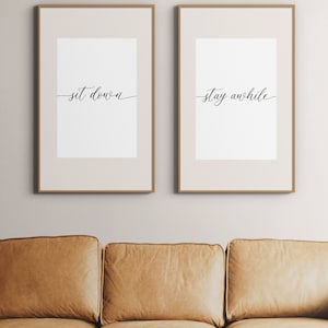 May include: Two framed art prints with the phrases "set down" and "stay awhile" in elegant cursive script. The frames are a light brown color. Below the prints is a tan leather couch and a small wooden coffee table.