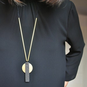 Peut inclure: Un collier avec un pendentif géométrique en or et noir, sur une longue chaîne fine. Le pendentif est composé de deux pièces : un disque rond et une barre rectangulaire.