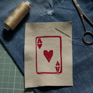 May include: A rectangular beige fabric patch featuring a red ink-stamped ace of hearts playing card design. The patch is positioned on blue denim fabric alongside a spool of tan thread, a silver sewing needle, and a pair of metal scissors.