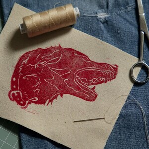 May include: A rectangular piece of beige canvas fabric featuring a red screen-printed graphic of a snarling wolf head. A spool of tan sewing thread and a metal sewing needle are placed on the fabric, alongside a pair of silver metal scissors.