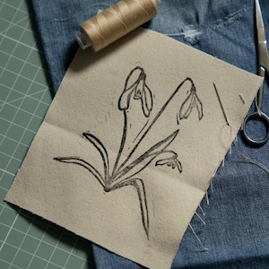 May include: A rectangular piece of beige fabric featuring a black ink block print of three snowdrop flowers. A spool of tan thread and a silver sewing needle are positioned nearby, resting on a piece of blue denim fabric.