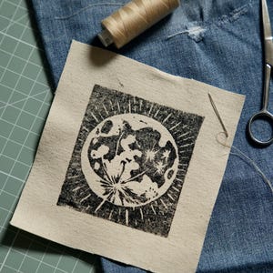 May include: A square piece of off-white fabric featuring a black ink block print of a full moon with radiating light rays. The fabric patch is accompanied by a spool of tan thread, a silver sewing needle, and a pair of metal scissors.